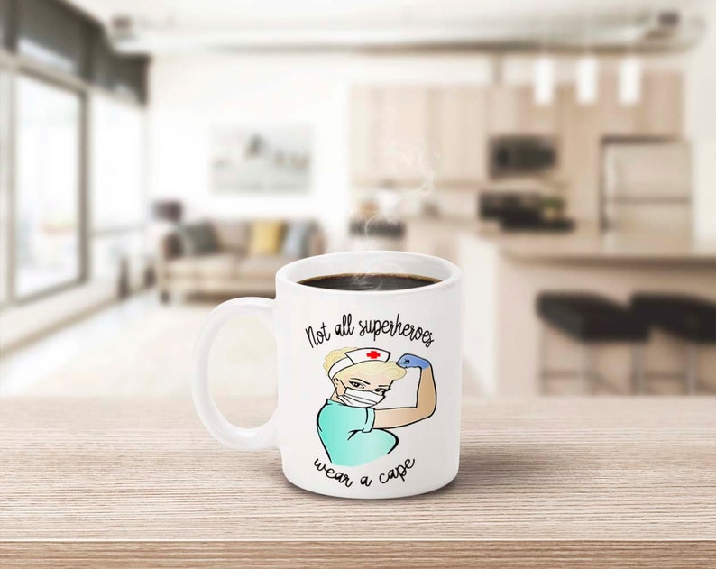 Peut inclure: Mug en c&eacute;ramique blanche remplie de caf&eacute; noir, avec une infirmi&egrave;re de dessin anim&eacute; montrant ses muscles. Le mug porte l'inscription "Not all superheroes wear a cape". L'infirmi&egrave;re porte un masque et une coiffe d'infirmi&egrave;re.