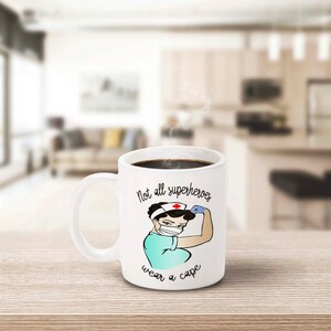 Peut inclure: Mug en c&eacute;ramique blanche avec l'inscription &laquo;&nbsp;Not all superheroes wear a cape&nbsp;&raquo;. La tasse pr&eacute;sente une illustration de dessin anim&eacute; d'une infirmi&egrave;re fl&eacute;chissant son bras. L'infirmi&egrave;re porte un masque facial, un bandeau avec une croix rouge et un uniforme turquoise.