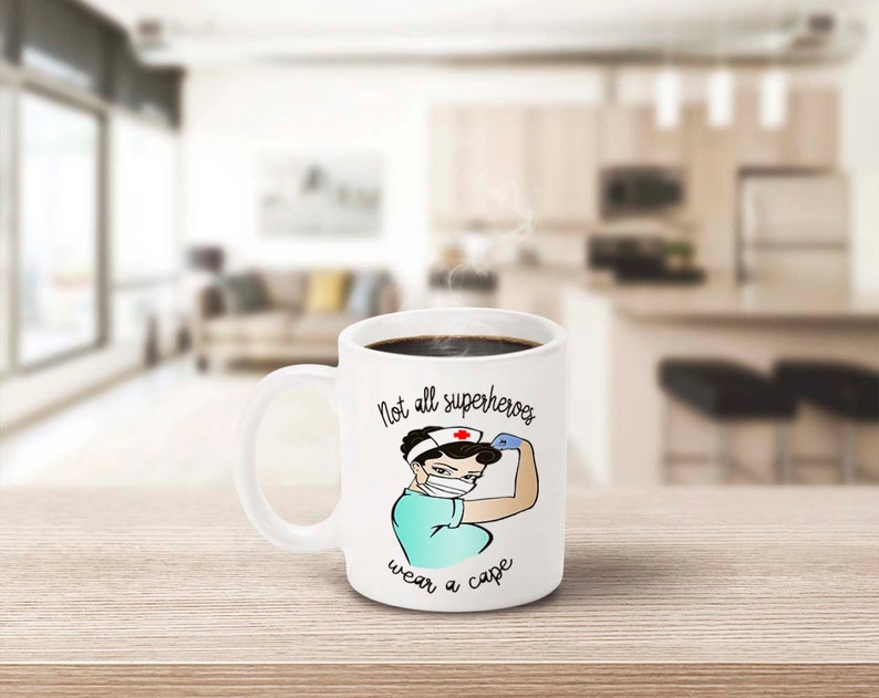 Peut inclure: Mug en c&eacute;ramique blanche avec l'inscription &laquo;&nbsp;Not all superheroes wear a cape&nbsp;&raquo;. La tasse pr&eacute;sente une illustration de dessin anim&eacute; d'une infirmi&egrave;re fl&eacute;chissant son bras. L'infirmi&egrave;re porte un masque facial, un bandeau avec une croix rouge et un uniforme turquoise.