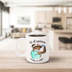 Peut inclure: Mug en c&eacute;ramique blanche avec une anse, remplie de caf&eacute; noir. La tasse pr&eacute;sente une illustration de dessin anim&eacute; d'une infirmi&egrave;re fl&eacute;chissant son bras, avec le texte "Not all superheroes wear a cape."