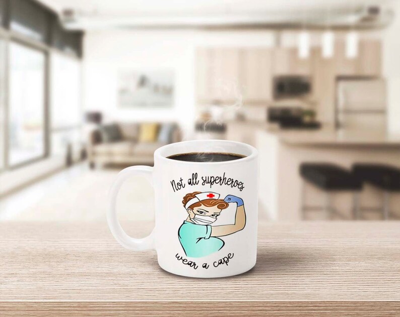 Peut inclure: Mug en c&eacute;ramique blanche avec une anse, rempli de caf&eacute; noir. La tasse pr&eacute;sente une illustration de dessin anim&eacute; d'une infirmi&egrave;re fl&eacute;chissant son bras, avec le texte "Not all superheroes wear a cape."