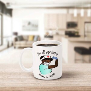 Peut inclure: Mug en c&eacute;ramique blanche remplie de caf&eacute; noir. La tasse pr&eacute;sente une illustration de dessin anim&eacute; d'une infirmi&egrave;re musclant son bras, avec le texte "Not all superheroes wear a cape". L'infirmi&egrave;re porte un masque et une blouse.