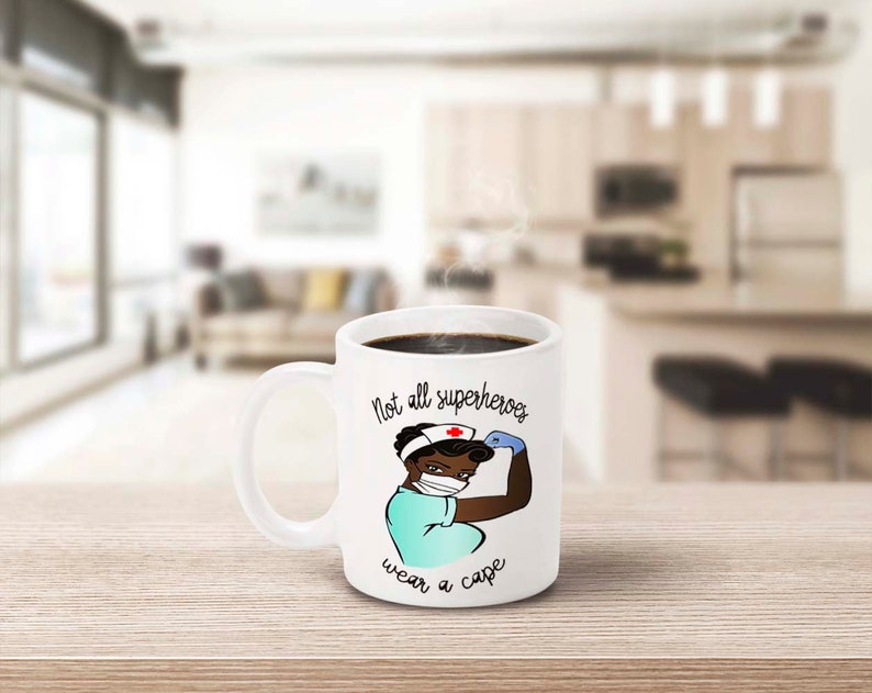 Peut inclure: Mug en c&eacute;ramique blanche remplie de caf&eacute; noir. La tasse pr&eacute;sente une illustration de dessin anim&eacute; d'une infirmi&egrave;re musclant son bras, avec le texte "Not all superheroes wear a cape". L'infirmi&egrave;re porte un masque et une blouse.