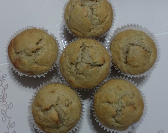 May include: Six banana nut muffins in white paper liners, arranged in a circle on a white surface. The muffins are golden brown and have a slightly domed top.