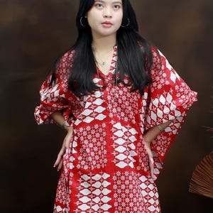 May include: A red and white patterned tunic with a V-neck and wide sleeves. The tunic features a repeating floral and geometric design. The model is wearing the tunic with blue jeans and silver hoop earrings.