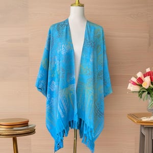 May include: A bright blue open-front kimono featuring a repeating pattern of tropical leaves in shades of green and gold. The garment has a loose, flowing silhouette with wide sleeves and a hemline finished with long, matching blue fringe tassels.