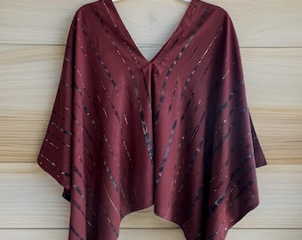 Pode incluir: Um poncho em tom bordô profundo com decote em V e uma série de riscas diagonais pretas em tie-dye que percorrem o tecido. A peça tem uma silhueta larga e fluida com bainhas pontiagudas, pendurada num cabide de plástico branco.