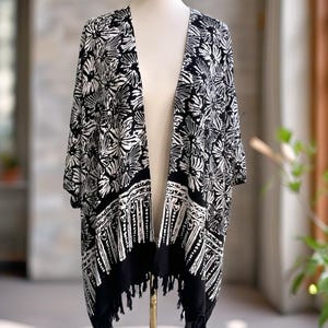 May include: A black and white open-front kimono featuring a bold floral pattern on the upper portion and a contrasting geometric border along the hem. The garment has short sleeves and is finished with a fringe detail at the bottom edge.