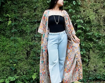 May include: A long, open-front kimono with a floral pattern in orange, yellow, and blue. The kimono is worn over a black tube top and light blue jeans. Black heeled sandals complete the outfit. The background is a lush green wall of foliage.