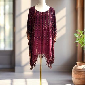 May include: A burgundy and dark brown tie-dye poncho featuring a round neckline, short sleeves, and a fringed hemline. The garment is displayed on a gold-toned mannequin stand, showcasing a vertical pattern of light and dark stripes.