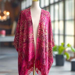 May include: A magenta open-front kimono featuring a gold-toned swirling floral pattern across the upper section and a geometric border design along the lower hem, finished with long, matching magenta fringe tassels at the bottom edge.