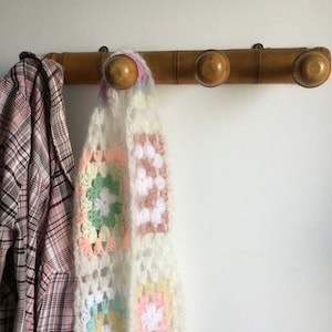 Peut inclure: Un porte-manteau en bambou avec quatre crochets, dont deux sont utilisés pour accrocher une écharpe en crochet avec des carrés de couleurs pastel et une chemise à carreaux.