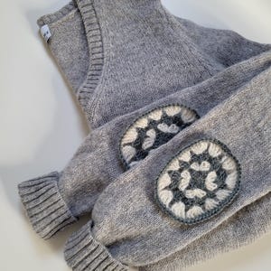 Peut inclure: Un pull en tricot gris avec des coudières. Les coudières présentent un motif au crochet blanc et gris. Le pull a un col en V et des poignets côtelés. Le pull est plié.