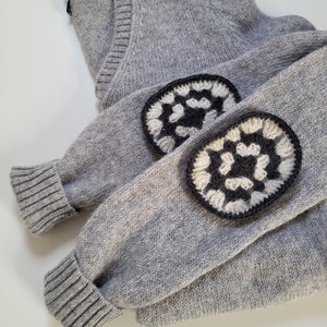 May include: A gray V-neck sweater with crocheted elbow patches. The elbow patches are a combination of black and white, with a floral design. The sweater is folded, showing the sleeves and the neckline.