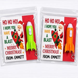 Peut inclure: Deux sacs cadeaux de Noël avec un porte-clés en forme de fusée et une carte. La carte dit : « Ho Ho Ho ! J'espère que vous vous amuserez + Joyeux Noël ! De : Emmett. » Un porte-clés est vert, l'autre est orange.