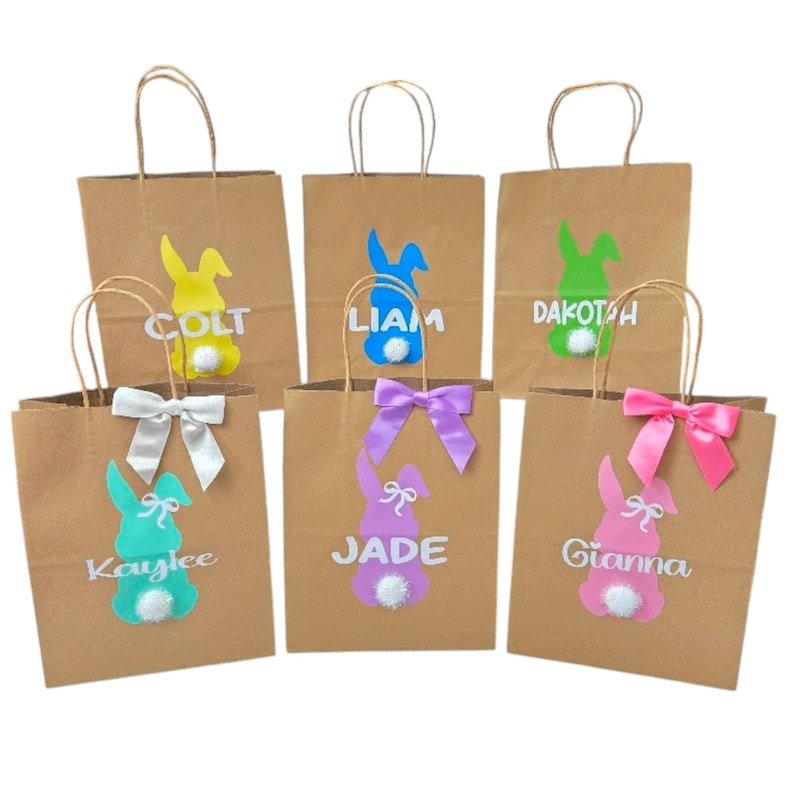 Puede incluir: Seis bolsas de regalo de papel marr&oacute;n con asas, cada una decorada con un dise&ntilde;o de conejito colorido y un nombre. Los conejitos son amarillos, azules, verdes, morados y rosas, con lazos a juego. Los nombres son en blanco.