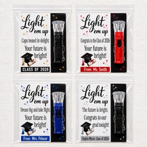 May include: Four individual graduation-themed gift sets in clear plastic bags, each containing a small flashlight and a card with the text Light 'em up, Your future is bright, and a graduation cap illustration, featuring black, red, and blue flashlights.