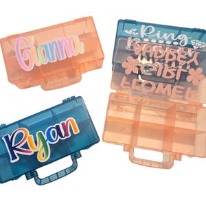 May include: Three clear plastic storage boxes with colorful names on the lids. The boxes are labeled "Gianna", "Ryan", and "Ring Girl Flower", and the last box has multiple compartments.