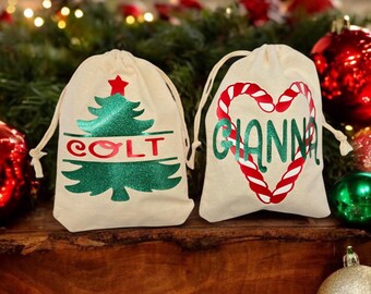 May include: Two personalized Christmas gift bags. One bag features a green Christmas tree with red glitter and the name "COLT" written in red. The other bag features a red and white candy cane heart with the name "GIANNA" written in green.