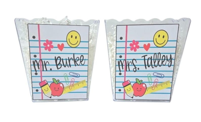 May include: Two clear, scalloped-edge containers filled with white shredded paper. Each container has a notepad design with a smiley face, hearts, and school supplies. One says "Mr. Burke" and the other says "Mrs. Talley".