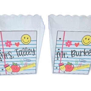 May include: Two white, scalloped-edge containers with notebook paper designs. Each features a smiley face, hearts, flowers, and school-themed graphics. One reads "Mrs. Talley" and the other "Mr. Burke".