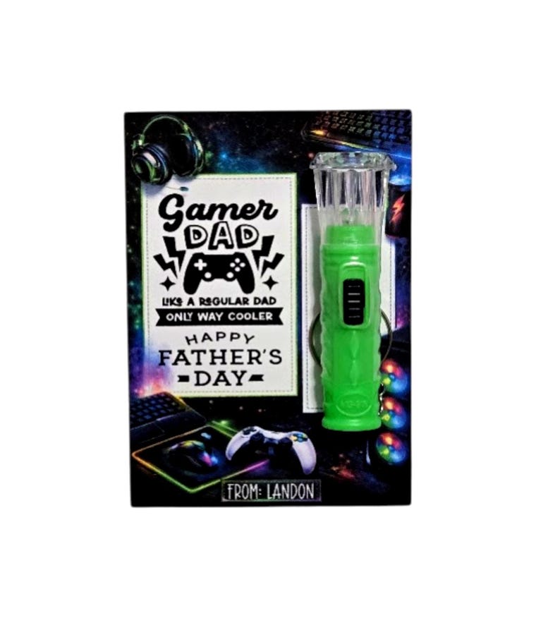 Pu&ograve; includere: Un biglietto per la festa del pap&agrave; a tema gaming. Il biglietto presenta il testo "Gamer Dad" e una grafica del controller. Un gadget verde illuminato &egrave; sulla destra. Il biglietto &egrave; decorato con accessori di gioco e il testo "From: Landon."