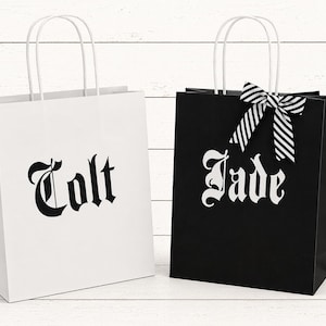 Puede incluir: Dos bolsas de regalo con asas blancas. Una bolsa es blanca con el nombre "Colt" en fuente gótica negra. La otra bolsa es negra con el nombre "Jade" en fuente gótica blanca y un lazo a rayas blanco y negro.