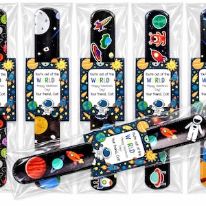May include: Six space-themed slap bracelets in clear plastic packaging. Each bracelet features colorful planets, rockets, and astronauts on a black background. Attached is a rectangular card with the text "You're out of this WORLD Happy Valentine's Day! Your friend, Colt."