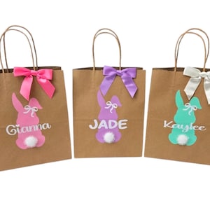 Puede incluir: Tres bolsas de regalo de papel marr&oacute;n con dise&ntilde;os de conejos. Cada bolsa tiene un lazo y una silueta de conejo de diferente color, con un nombre impreso debajo. Las bolsas est&aacute;n decoradas con lazos rosas, morados y blancos.