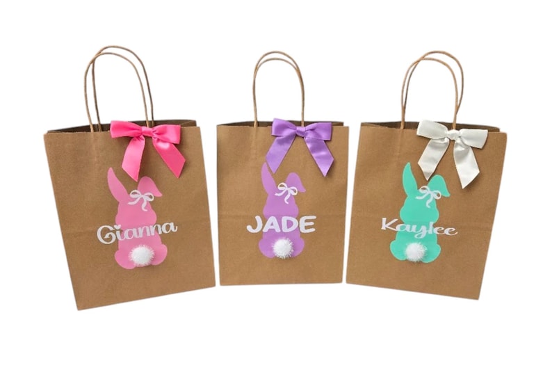 Puede incluir: Tres bolsas de regalo de papel marr&oacute;n con dise&ntilde;os de conejos. Cada bolsa tiene un lazo y una silueta de conejo de diferente color, con un nombre impreso debajo. Las bolsas est&aacute;n decoradas con lazos rosas, morados y blancos.