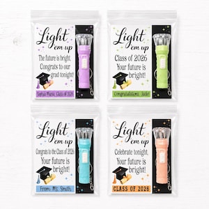 May include: Four graduation-themed party favors in clear plastic bags. Each bag contains a small flashlight in purple, green, blue, or peach. Text on the bags reads "Light 'em up" and congratulatory messages for the Class of 2026.