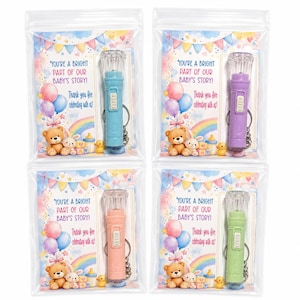 May include: Four small, colorful flashlights in clear plastic bags. Each flashlight is a different color: blue, purple, pink, and green. The bags have a card with text and illustrations of balloons, a rainbow, and teddy bears. The text reads: "YOU'RE A BRIGHT PART OF OUR BABY'S STORY! Thank you for celebrating with us!"