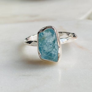 May include: A silver ring featuring a light blue aquamarine gemstone. The ring has a hammered texture and the gemstone is set in a bezel setting.