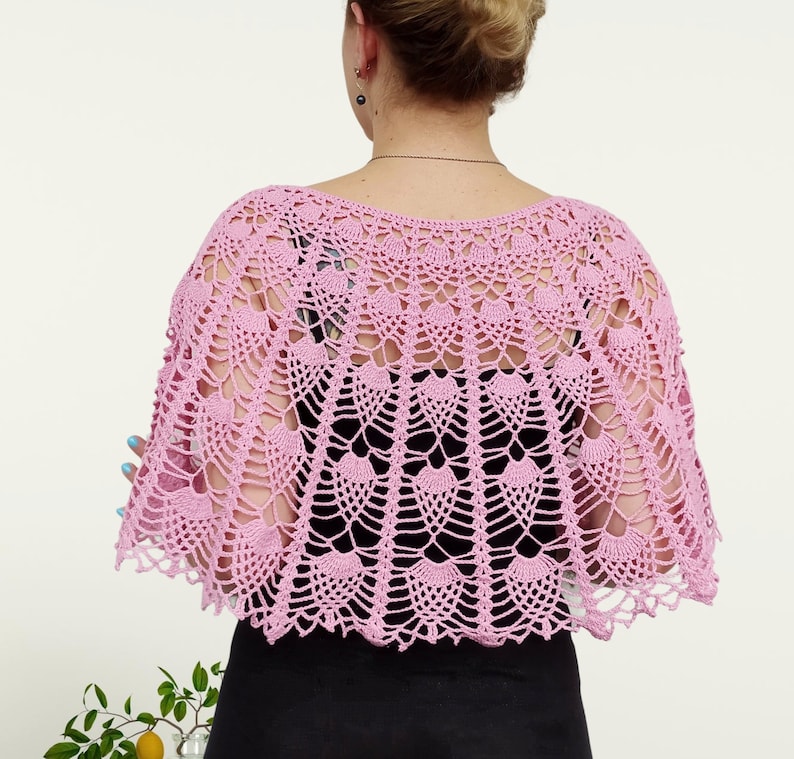 Op de afbeelding: Een lichtroze gehaakte sjaal met een ingewikkeld opengewerkt patroon met herhalende waaiervormige motieven. Het kledingstuk is over de schouders gedrapeerd en toont een geschulpte rand en een delicate, luchtige constructie van fijn garen.