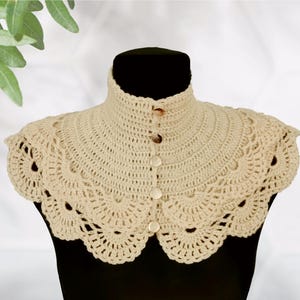 May include: A cream-colored crochet collar featuring a high neck and a scalloped edge design. The garment is fastened down the center front with five small, round, light-colored buttons. The intricate yarn work creates a layered, textured pattern.