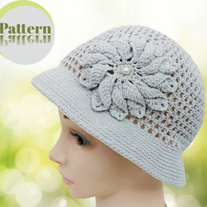 Puede incluir: Un sombrero de crochet gris con un diseño de flor y un acento de perla. El sombrero tiene un ala ancha y un delicado patrón de calado.