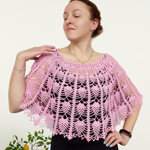 Op de afbeelding: Een lichtroze gehaakte poncho met een ingewikkeld opengewerkt patroon met herhalende waaiervormige motieven. Het kledingstuk heeft een brede, ronde halslijn en een geschulpte rand die over de schouders valt, gedragen over een effen zwart topje.