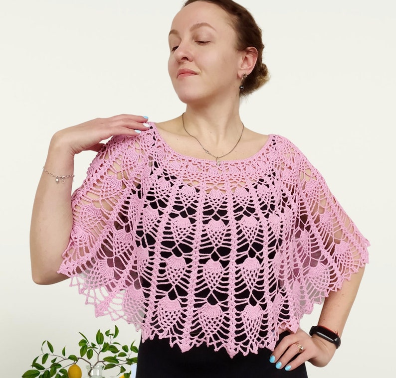 Op de afbeelding: Een lichtroze gehaakte poncho met een ingewikkeld opengewerkt patroon met herhalende waaiervormige motieven. Het kledingstuk heeft een brede, ronde halslijn en een geschulpte rand die over de schouders valt, gedragen over een effen zwart topje.