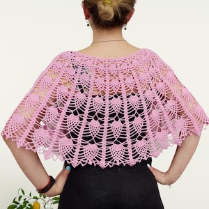 Op de afbeelding: Een lichtroze gehaakte capelet met een ingewikkeld, opengewerkt waaierpatroon. Het kledingstuk heeft een brede, geschulpte halslijn en een uitlopend, halfrond silhouet dat over de schouders valt en gedetailleerd handwerk laat zien.