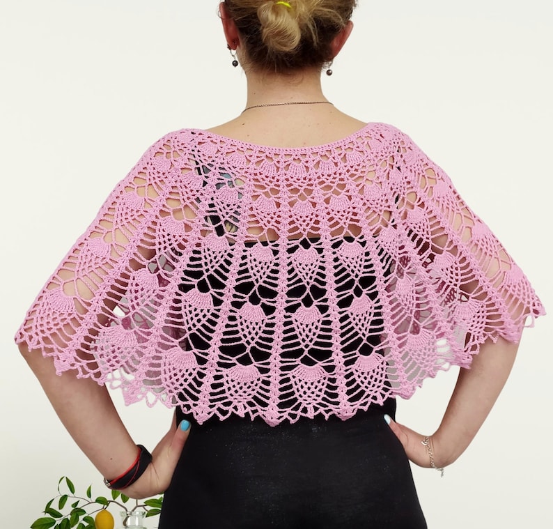 Op de afbeelding: Een lichtroze gehaakte capelet met een ingewikkeld, opengewerkt waaierpatroon. Het kledingstuk heeft een brede, geschulpte halslijn en een uitlopend, halfrond silhouet dat over de schouders valt en gedetailleerd handwerk laat zien.