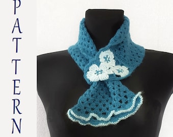 Può includere: Una sciarpa all'uncinetto blu turchese con fiori all'uncinetto bianchi. La sciarpa ha un bordo a festoni.
