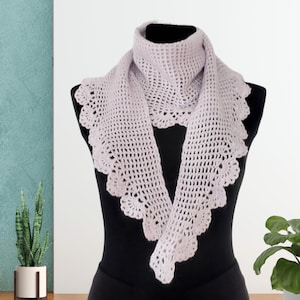 May include: A light purple crochet scarf with a scalloped edge is displayed on a black mannequin. The scarf has an open weave design and is draped around the mannequin's neck and shoulders. The background is a light blue wall with a potted plant.