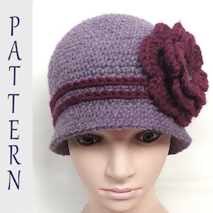 Puede incluir: Un gorro cloche de crochet morado con un adorno de flor burdeos. El gorro tiene un ala ancha y una banda doble de hilo burdeos alrededor de la corona.