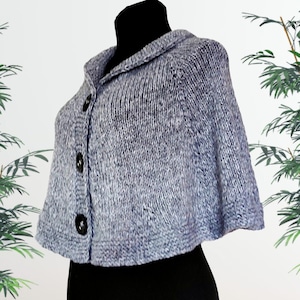 May include: A light blue, heathered knit capelet featuring a folded collar and a front placket secured with three large, dark, circular buttons. The garment has a textured, chunky knit construction with a ribbed hem and a cropped silhouette.