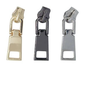 Puede incluir: Tres tiradores de cremallera en dorado, negro y plateado. Cada tirador tiene una forma rectangular con una pequeña abertura rectangular en la parte inferior.