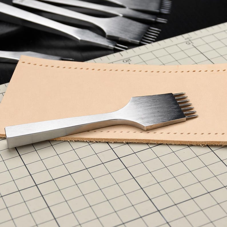 May include: A stainless steel leather stitching chisel with multiple prongs, resting on a piece of tan leather. Other similar tools are visible in the background, suggesting a leathercrafting theme. The tools are on a grid-lined surface.
