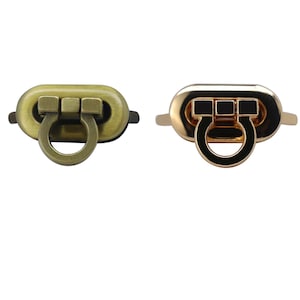 Könnte beinhalten: Zwei Metallschließen, eine goldfarben und eine antik-messingfarben, mit einem U-förmigen Ring. Die Schließen haben ein rechteckiges Design mit drei erhabenen Quadraten.