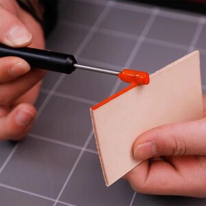 Peut inclure: Une personne utilise un outil pour appliquer de la peinture orange sur le bord d'un morceau de cuir.