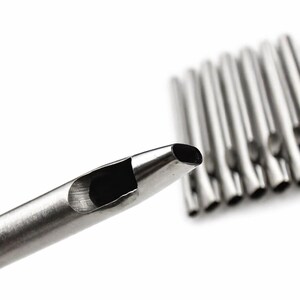 May include: Close-up of a silver-colored leathercraft punch tool with a hollow, oval-shaped cutting end. Several similar tools are blurred in the background, all against a white backdrop. The tools are designed for creating holes in leather and similar materials.