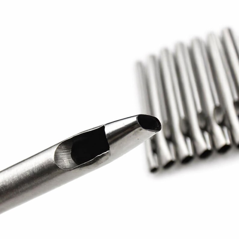 May include: Close-up of a silver-colored leathercraft punch tool with a hollow, oval-shaped cutting end. Several similar tools are blurred in the background, all against a white backdrop. The tools are designed for creating holes in leather and similar materials.